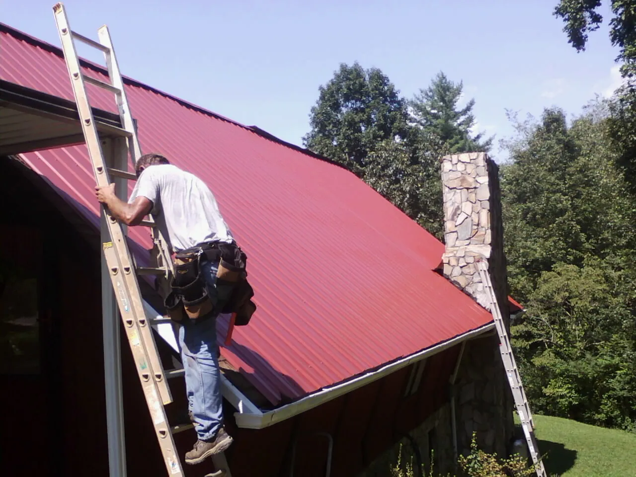 Roofer on a ladder beside a red metal roof and stone chimney — Soffit & Fascia Repair by Roofing Brothers in Mill Run